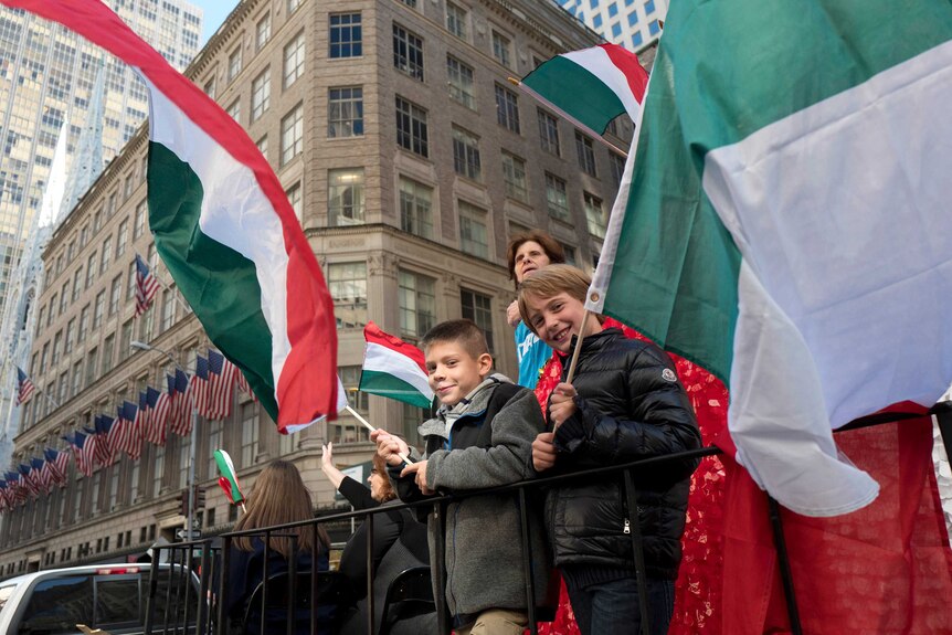 Two boys are waving large Italian flags while standing on a float. An office building with American flags is in the background.