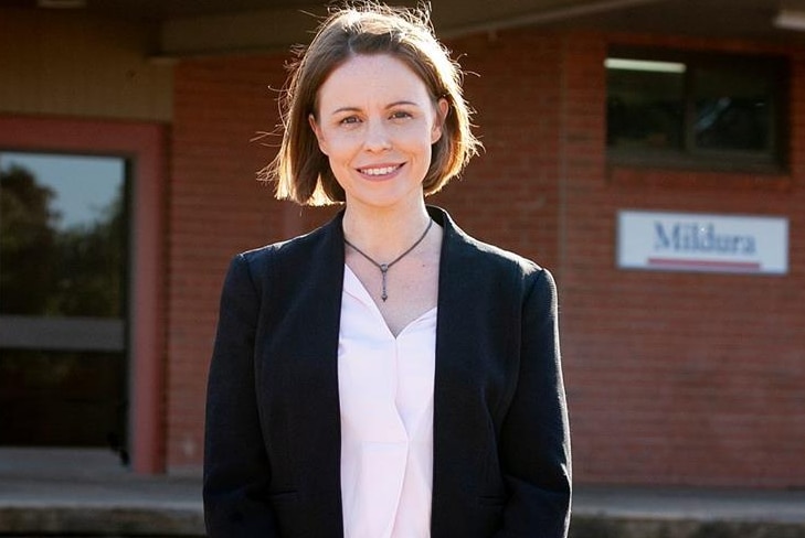 A young woman in a black jacket smiles at the camera while standing in front of Mildura train station.