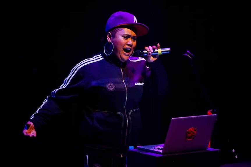A black woman with a mic singing in front of a laptop on stage.