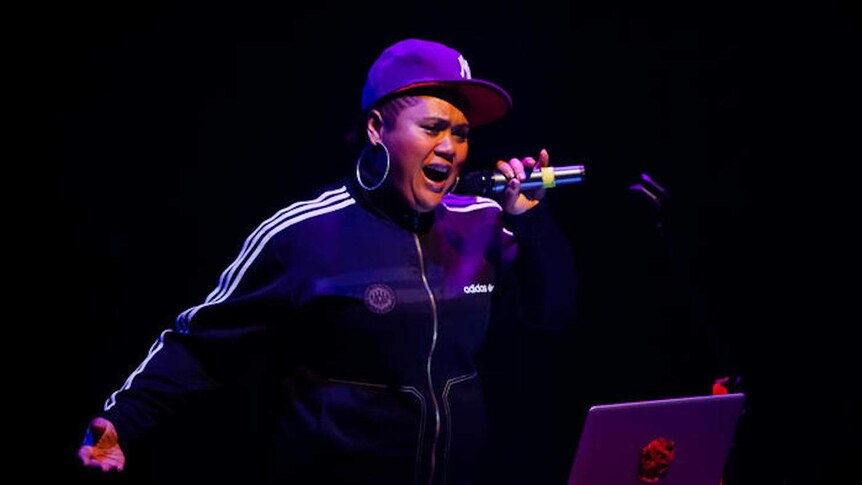 A black woman with a mic singing in front of a laptop on stage.