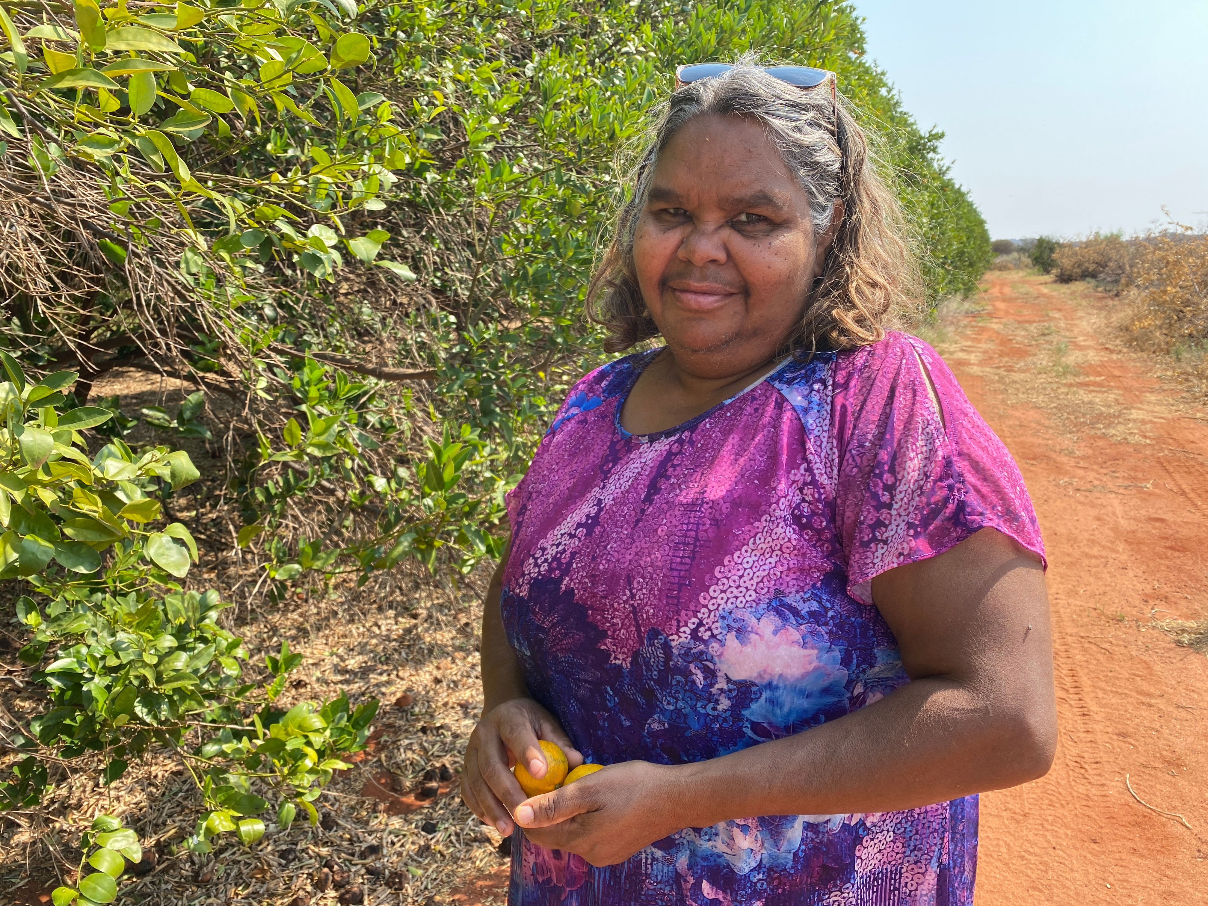 An Aboriginal woman wearing a bright purple shirt, stands in front of overgrown orange trees on red sandy soil.