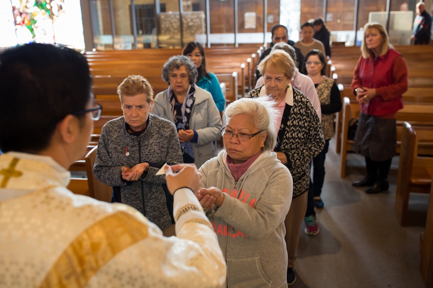 A congregation of older parishioners line up in the church aisle, hands cupped to receive communion.