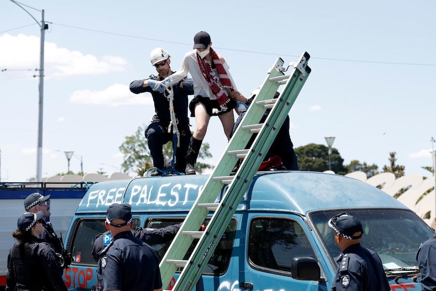A woman on top of a blue van is arrested by a police officer.