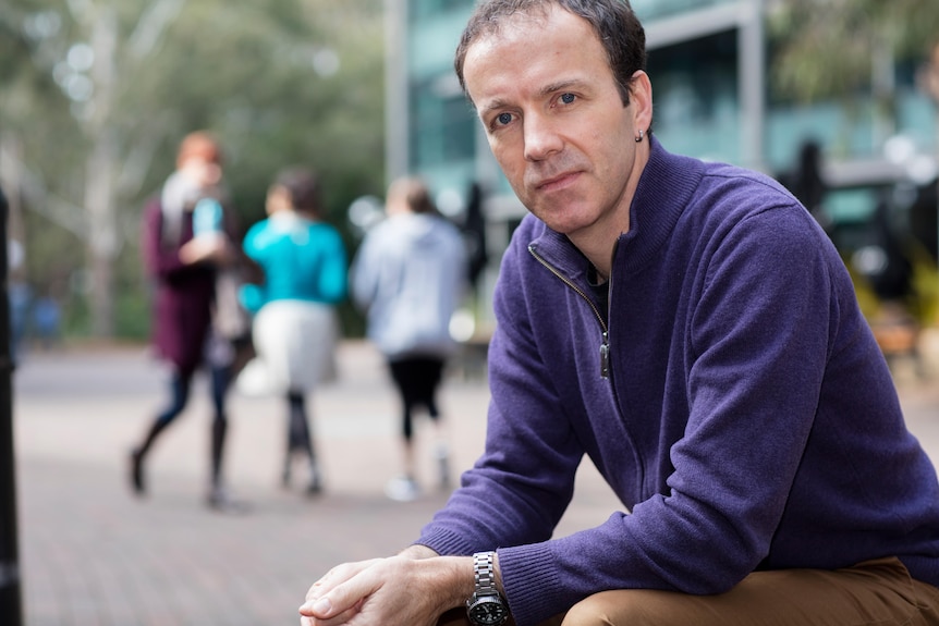 Michael Flood wears a purple jumper and a serious expression in an outdoor setting