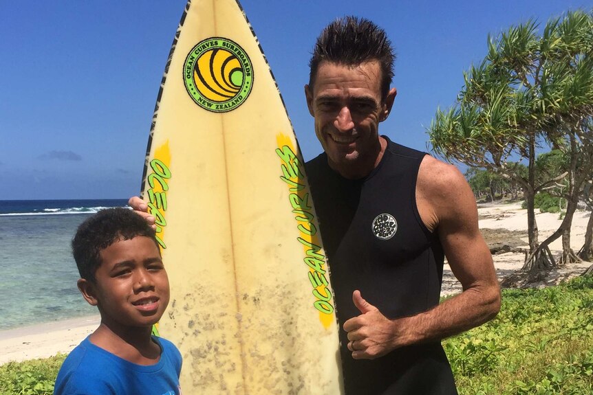 Profile photo of a young boy and a man holding a surfboard on a beach.
