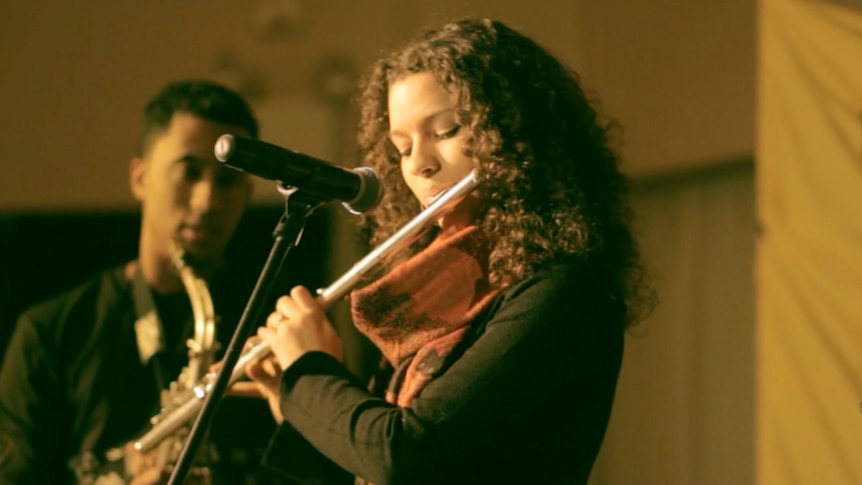 Elena Pinderhughes playing her flute into a microphone; she's wearing a red scarf and a green jumper