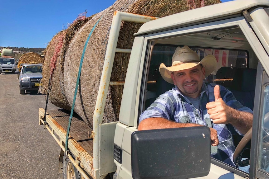 A farmer wearing a straw cowboy hat gives the thumbs up from his ute as he drives away with two big bales of hay.