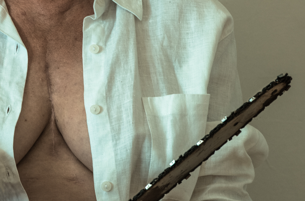 A woman holding a chainsaw, wearing a shirt open wide enough to show the heart operation scar between her breasts.