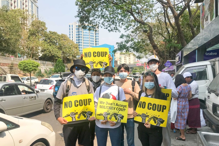 A group of people holding signs with "no coup" written on them on a street lined with trees.