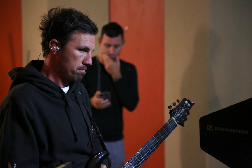 Two men stand in a room, the man in foreground appears annoyed