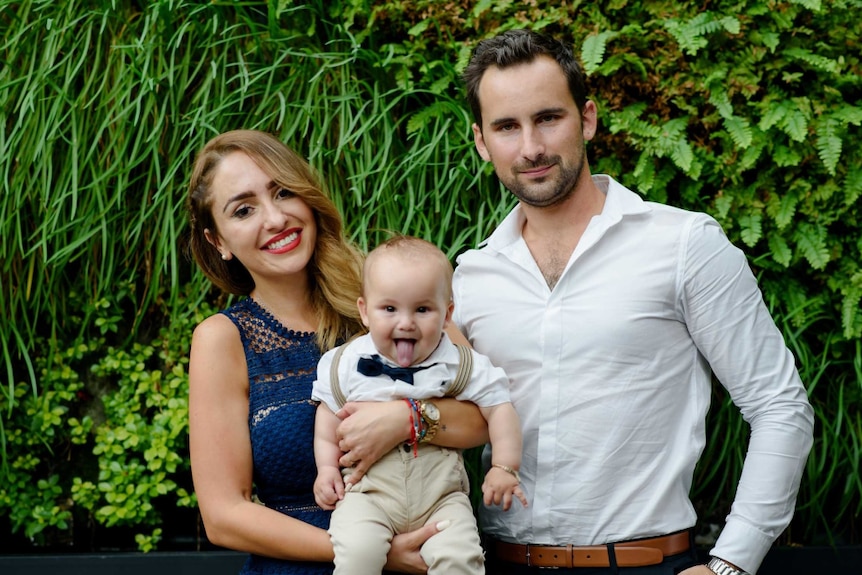 A man stands with a woman who holds a baby in front of leafy foliage.