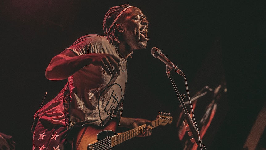 Bloc Party frontman Kele Okereke yells into the mic live on stage, holding his guitar and wearing a t-shirt and US flag shorts