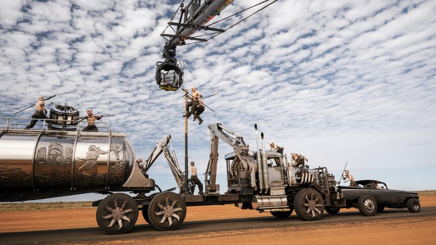 On set of Furiosa A Mad Max Saga, a silver truck rams a car and many warriors scamper on, a camera capturing the action