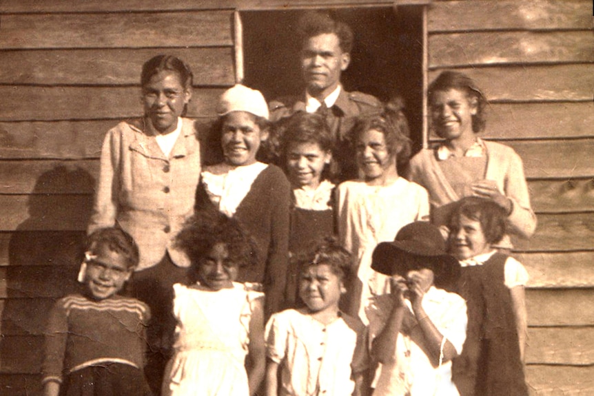 Group photo of 14 school children and teacher standing at the doorway of a wooden school building in the 1940s.