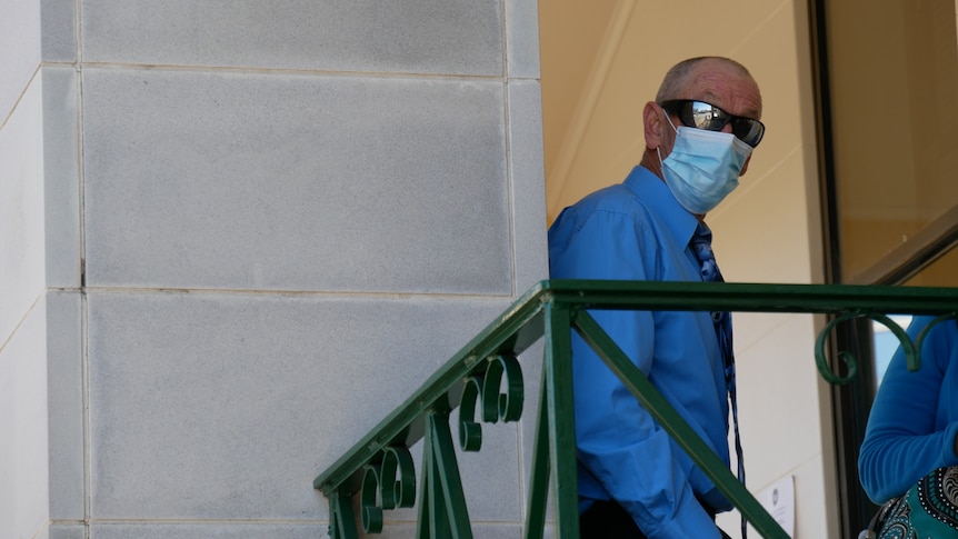  A bald man, wearing sunglasses, blue shirt, tie and blue surgical face mask stands behind a metal balustrade, looks at camera.