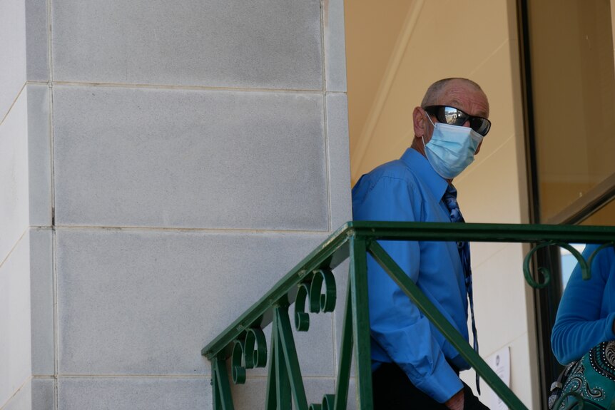  A bald man, wearing sunglasses, blue shirt, tie and blue surgical face mask stands behind a metal balustrade, looks at camera.