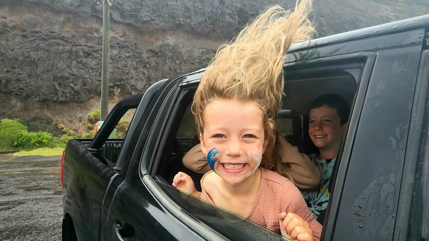 a little girl leans out the window of a parked car smiling, it's very windy and her blonde hair is blowing straight up