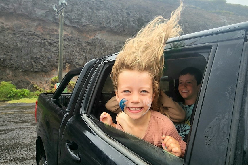 a little girl leans out the window of a parked car smiling, it's very windy and her blonde hair is blowing straight up