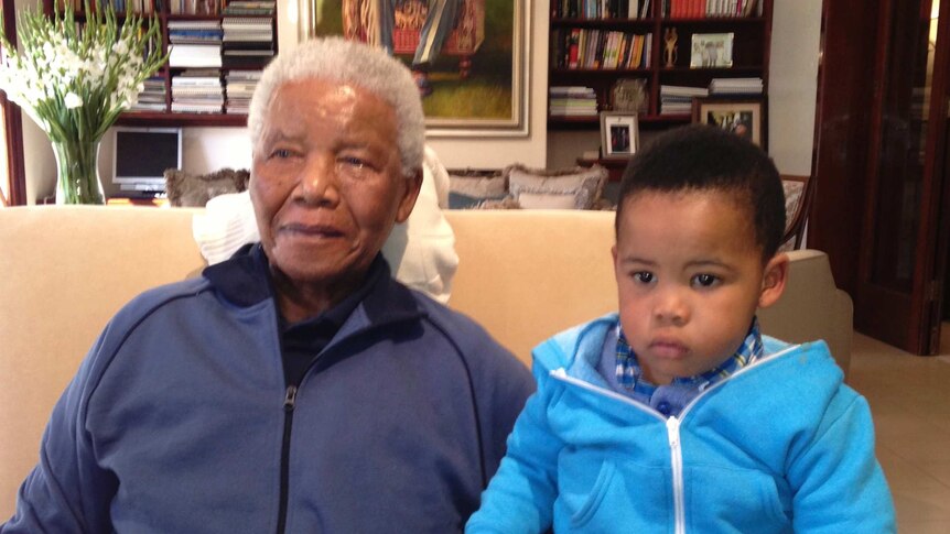 In a room lined with bookshelves, a smiling older man and a neutral-faced young child sit on a couch holding hands.