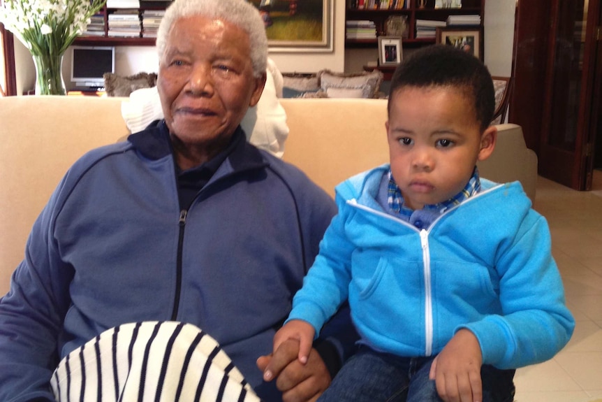 In a room lined with bookshelves, a smiling older man and a neutral-faced young child sit on a couch holding hands.