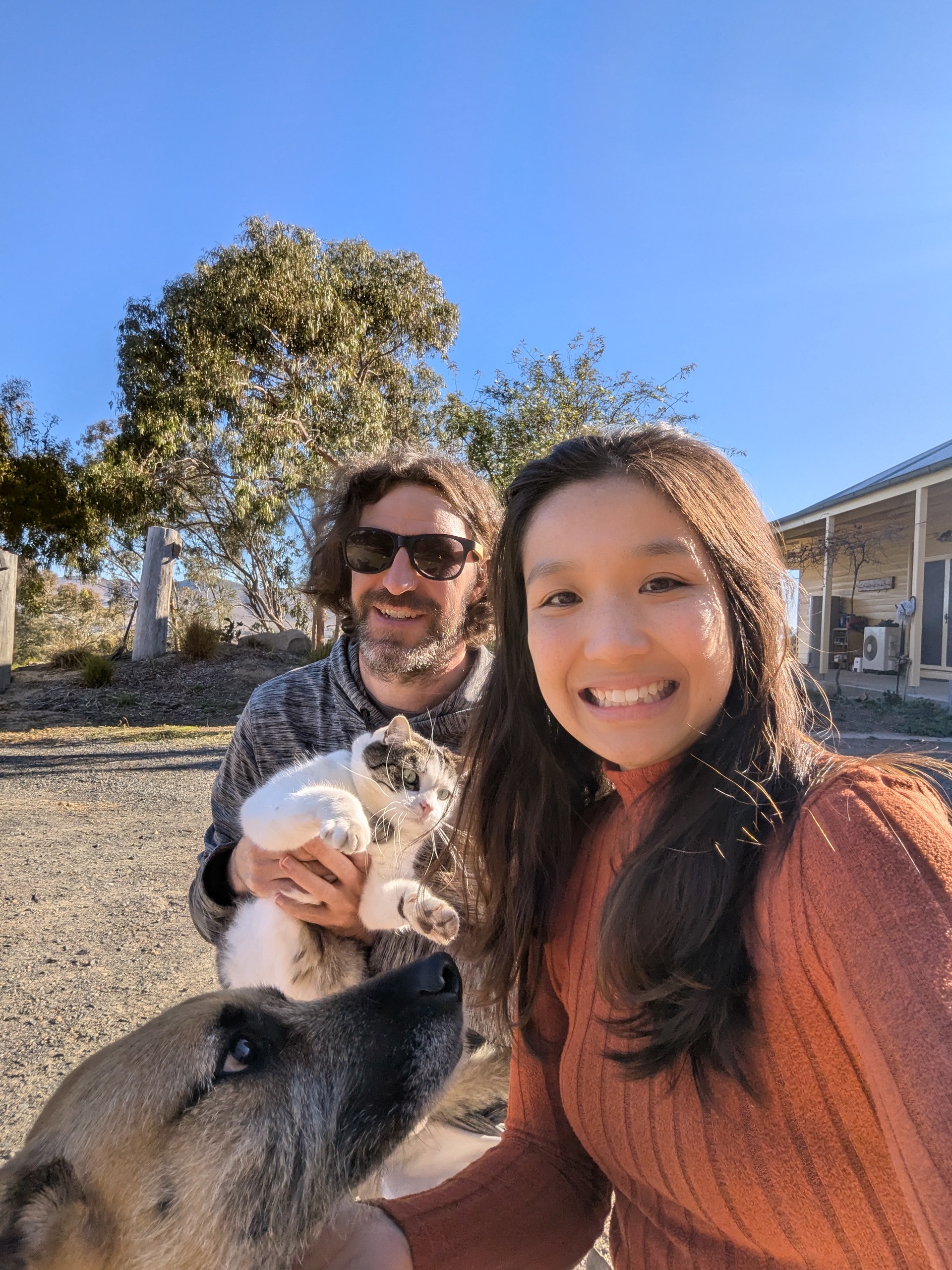 Winnie and Nelson pose for a selfie with their cat and dog. There is green grass and a tree behind them.