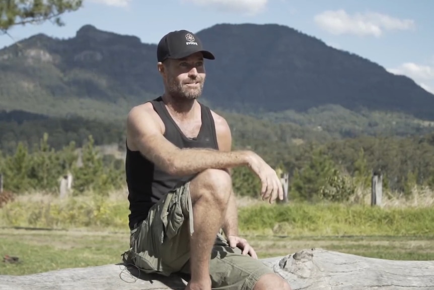 A man in a black singlet and hat sitting on a rock with mountains behind