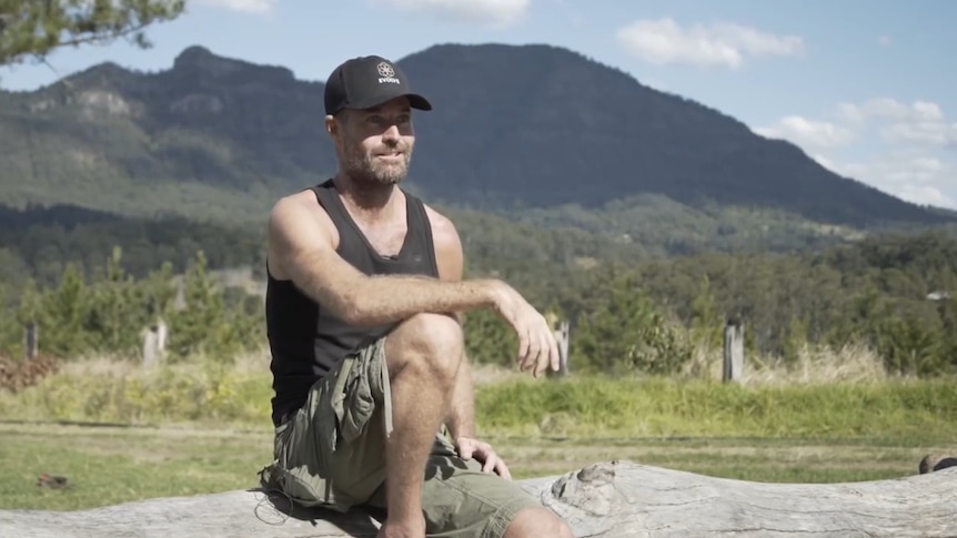 A man in a black singlet and hat sitting on a rock with mountains behind