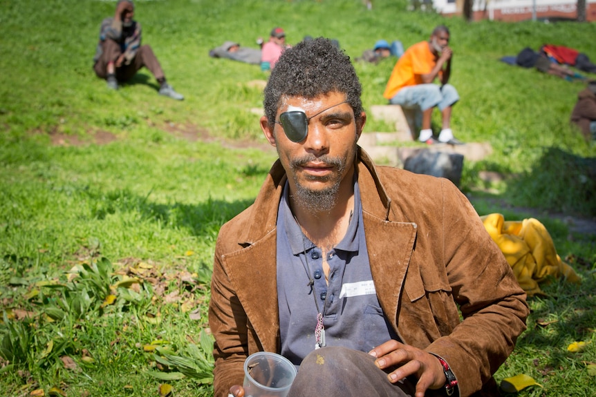 Man wearing eyepatch and brown jacket sits on grass.
