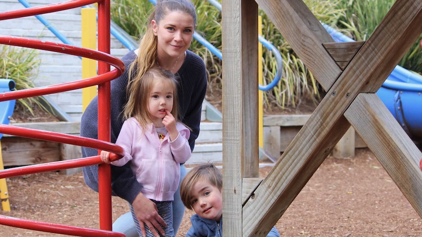 Oliver Tsuha-Brown kneels down in the middle of a playground with his sister and mother behind him.