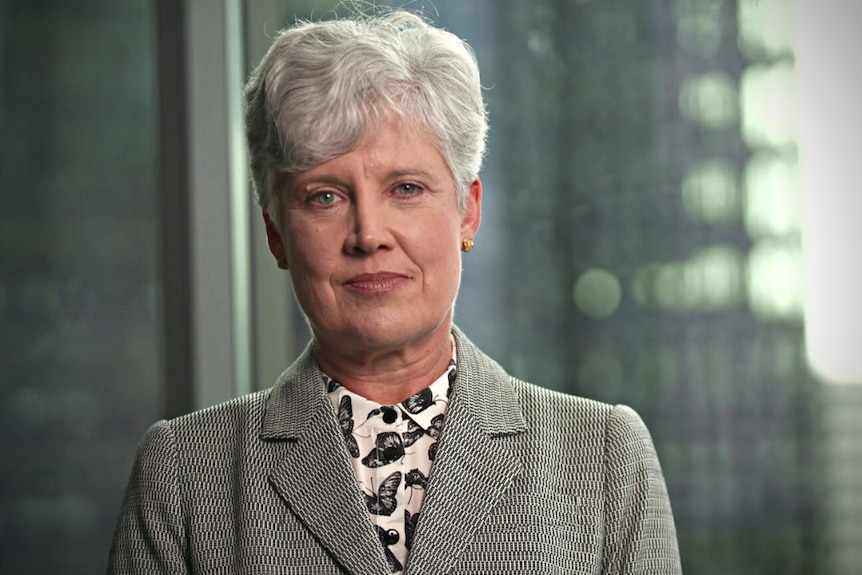 Woman with short grey hair wearing a black and white printed top and a grey jacket, sitting in an office.