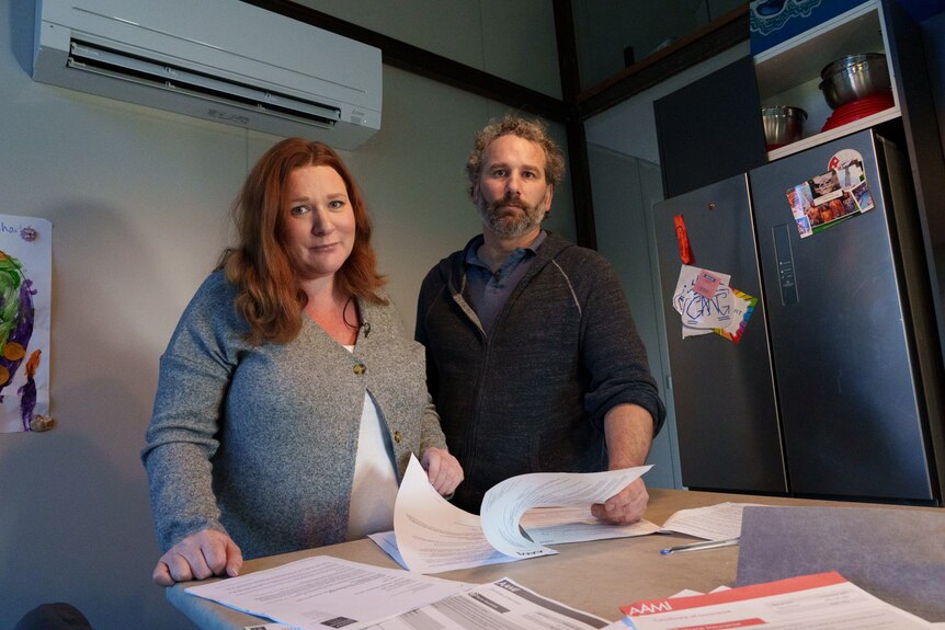 Verity and Justin lean on their kitchen bench surrounded by documents looking down at the camera.