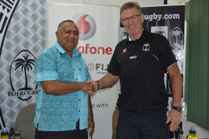 Francis Kean shakes hand with John McKeen against a Fiji Rugby and rugby.com banner
