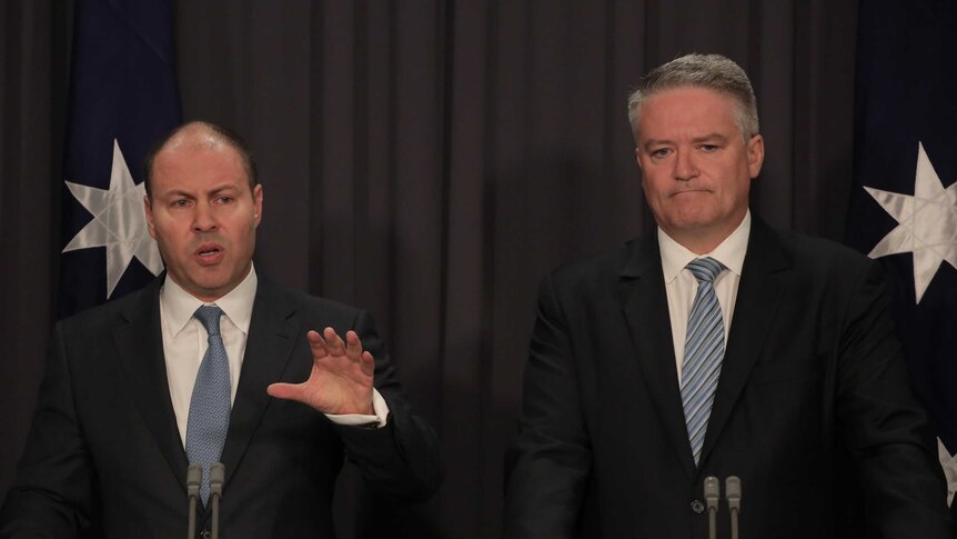 Josh Frydenberg and Mathias Cormann stand in front of lecterns with flags behind them
