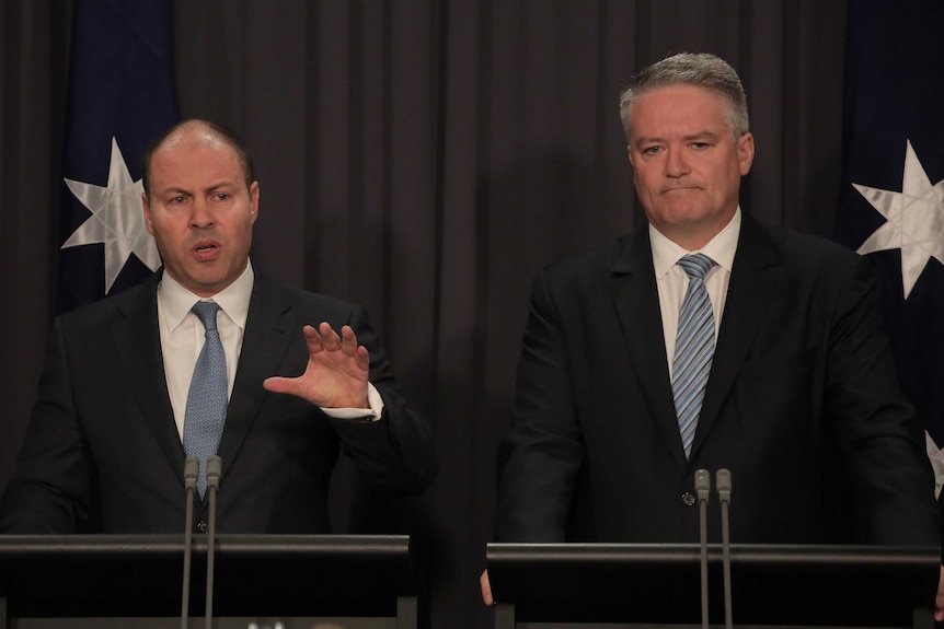 Josh Frydenberg and Mathias Cormann stand in front of lecterns with flags behind them