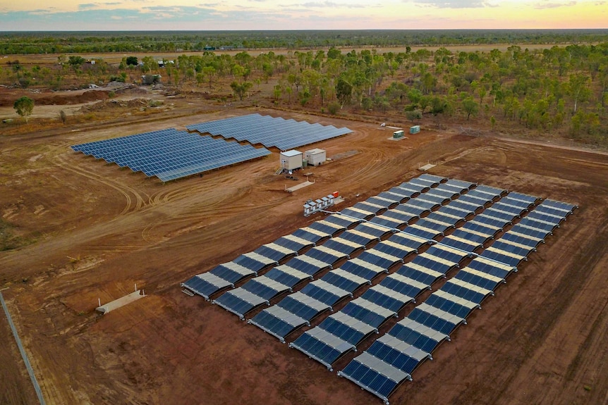A drone shot of rows of shining solar panels on red dirt with a flat treed landscape behind.
