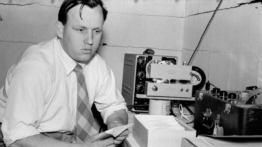 Black and white photo of a man in front of a desk with an oscilloscope, camera, and tape recorder.