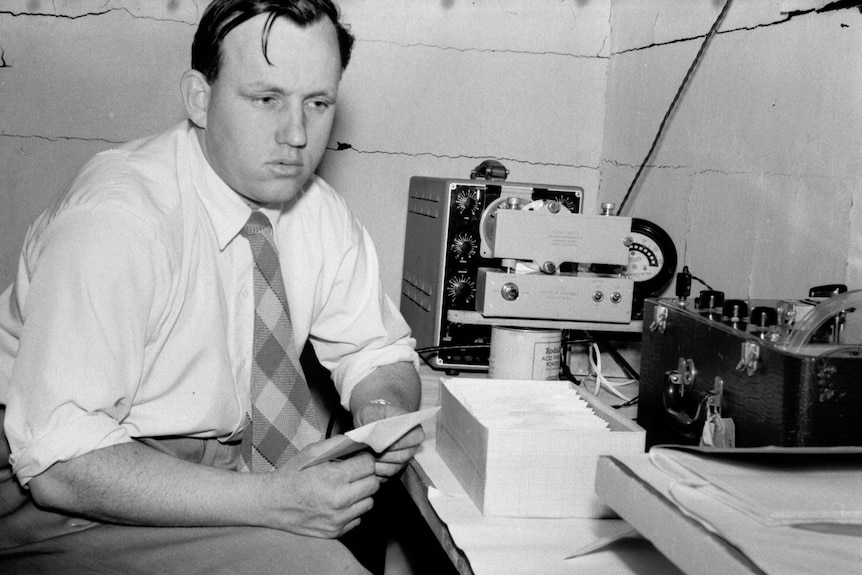 Black and white photo of a man in front of a desk with an oscilloscope, camera, and tape recorder.