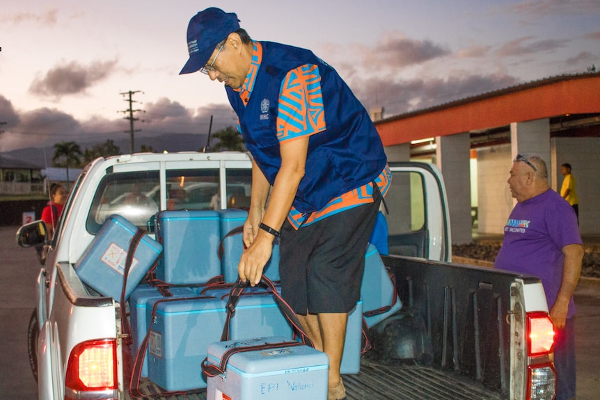 Man in back of ute picks up blue Styrofoam box while man in purple shirt watches. 