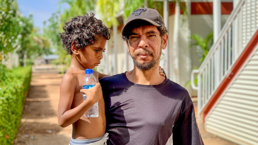 An Indigenous man wearing a black cap and T-shirt holds a small child, who is clutching a water bottle, outside a building