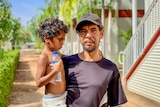 An Indigenous man wearing a black cap and T-shirt holds a small child, who is clutching a water bottle, outside a building