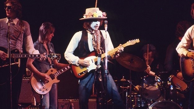 Bob Dyan with white paint on his face, wearing a straw hat decorated with flowers, plays a guitar on stage