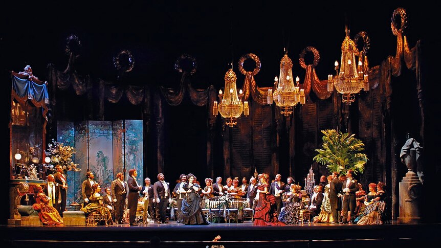 Revelers on stage under extravagant chandeliers in a production of Verdi's opera La Traviata for Florida Grand Opera.