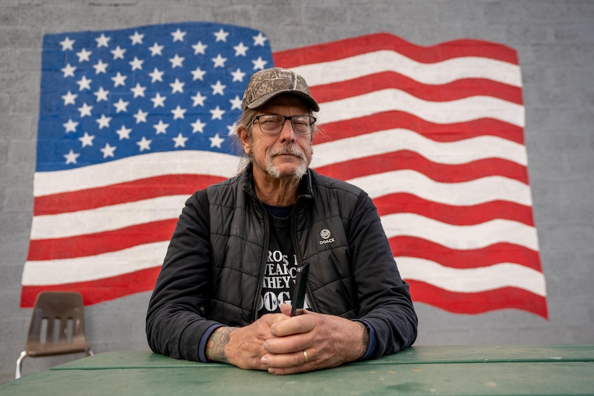 A man wearing a camo hat and holding a blazer sitting at a bench with a big american flag mural behind him