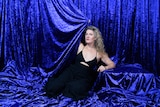 Woman with curly blonde hair and blue eyes sits in a blue velvet curtain and pulls it across her.