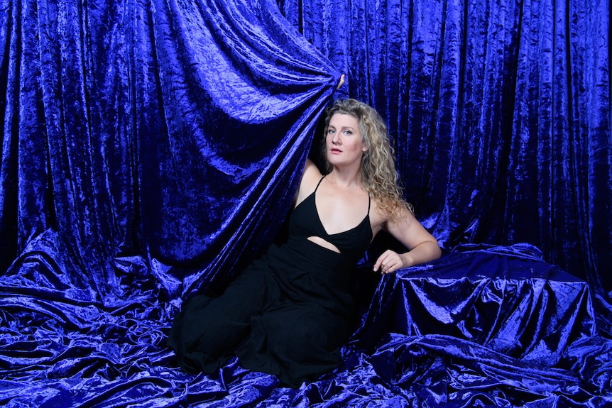 Woman with curly blonde hair and blue eyes sits in a blue velvet curtain and pulls it across her.