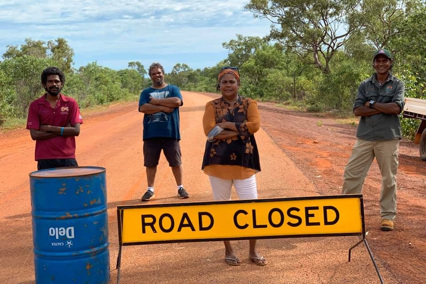 Four Indigenous people stand in a line across a road with a road closure sign