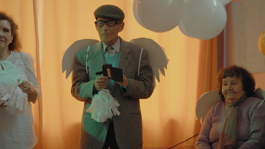 An elderly man, the spy Sergio Chamy, wearing angel wings standing in between two elderly women in documentary The Mole Agent