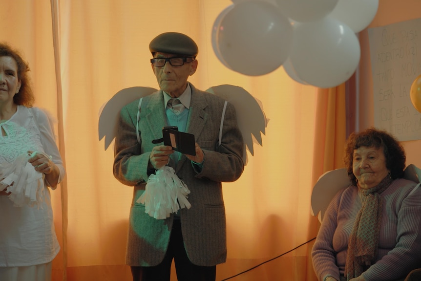 An elderly man, the spy Sergio Chamy, wearing angel wings standing in between two elderly women in documentary The Mole Agent