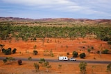 A four-wheel-drive towing a caravan along a bitumen road, captured by drone, surrounded by red dirt and small green shrubs.