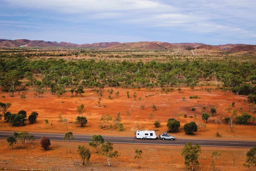 A four-wheel-drive towing a caravan along a bitumen road, captured by drone, surrounded by red dirt and small green shrubs.